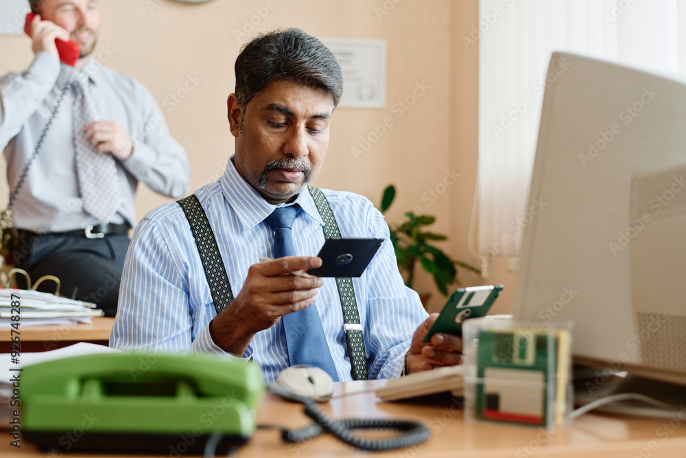 Poster middle-aged man sitting at desk handling two smartphones while colleague is talking in background du