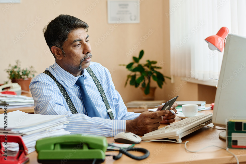 Poster businessman sitting at office desk holding smartphone in one hand while looking thoughtfully at comp