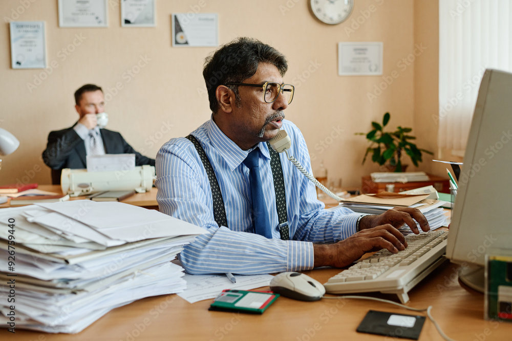 Poster Portrait of businessman typing on keyboard and using telephone in busy office environment with colleague in background drinking coffee