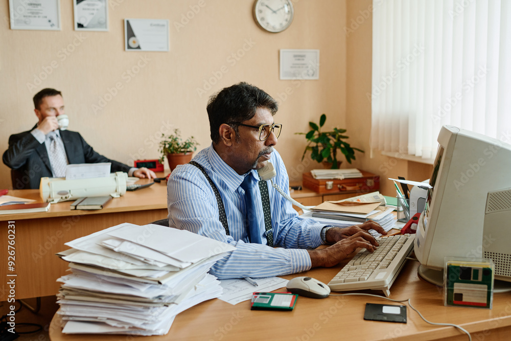 Poster Man wearing glasses talking on phone and typing on computer with office equipment and documents on desk while colleague in background drinks coffee and observes