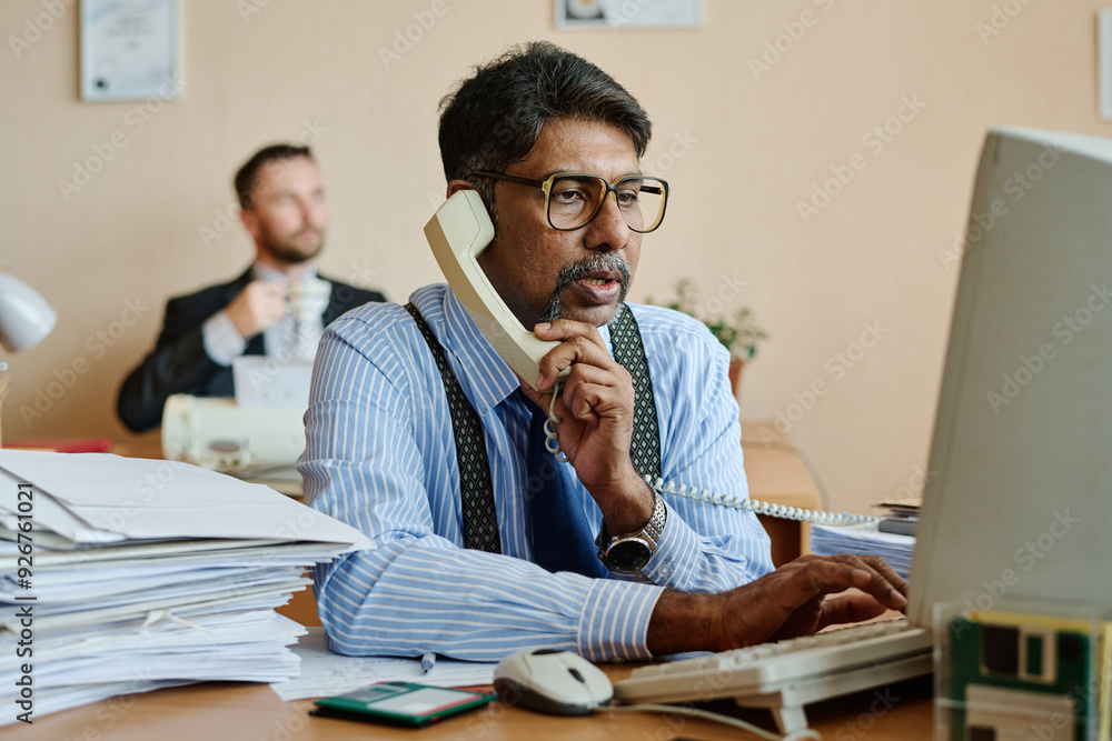 Leinwandbilder Caucasian businessman handling phone call while working at desk in office setting with colleague in background engaged in work activities creating professional atmosphere