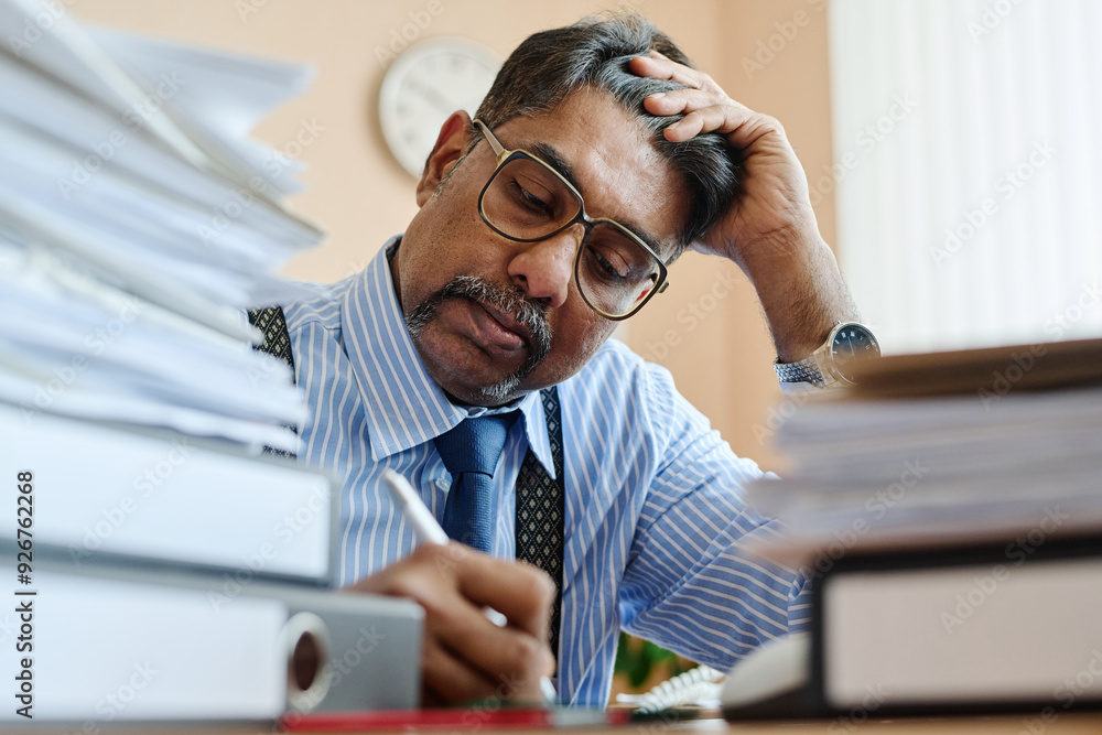 Poster Man focusing on work at cluttered office desk while writing on notebook. Desk covered with stacks of papers and books indicating a busy work environment