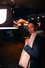 Beautiful girl in winter clothes stands near a lantern at night against the backdrop of a Christmas market. A young woman walks at night at a deserted New Year's fair and erases on a lamp