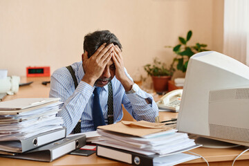 Man sitting at desk with head in hands, showing stress from piles of documents and paperwork...