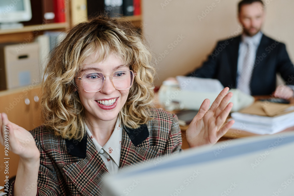 Poster Smiling woman gesturing enthusiastically while sitting at desk in office environment with colleague working in background, capturing excitement and positive vibes