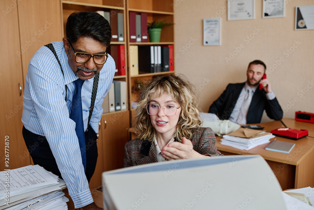 Poster Two colleagues discussing a project while another coworker is on a phone call in background. Office setting features documents, a computer, and organizing shelves