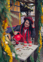 A young European woman in a red hat and scarf pours tea from a red thermos into cups at a Christmas...