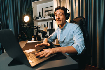 Fototapeta premium Host channel smart broadcaster talking on live social media application searching on laptop, broadcasting streamer wearing headphone using mic at modern home office studio in warm lighting. Pecuniary.
