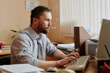 Man sitting at desk in office, smoking cigarette while typing on keyboard. Papers and office...