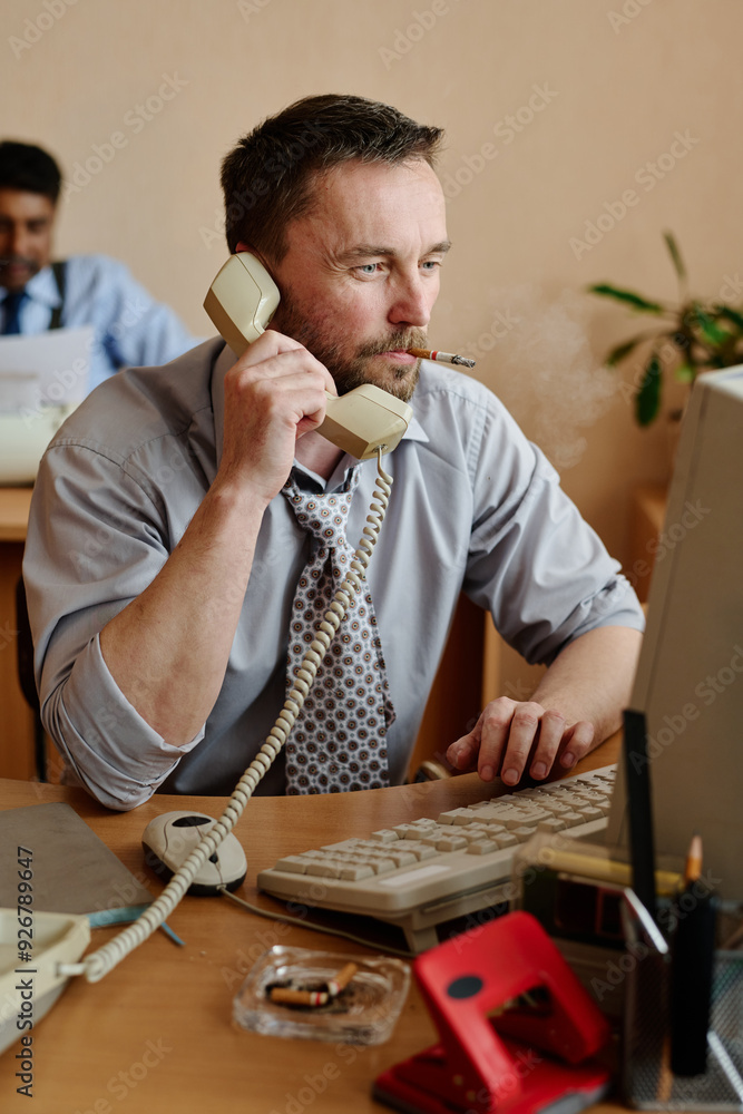 Fototapete Man sitting at office desk holding phone receiver and smoking with computer monitor in front. Colleague blurred in background working at another desk, creating a busy workspace vibe