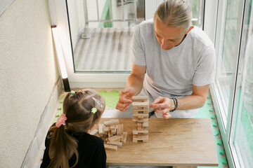 Child and an adult focus on building wooden tower game together at table indoors. Father and daughter enjoying free time. Quality family communication and informal education