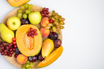 A plate filled with a variety of fruits, melon, apple, pear, grapes and peach on a light background. Top view, horizontal, free copy space.