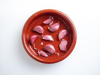 Close-up of garlic cloves placed in a rustic red clay bowl against a clean white background. The...