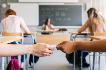 Young teenage boy giving cheat sheet to classmate during examination in classroom. College students cheating on an exam. Focus on students hands. Education lifestyle concept.