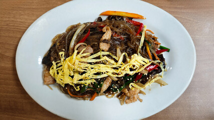Delicious Plate of Korean Japchae with Stir-Fried Glass Noodles, Vegetables, meat, and Egg Garnish on a White Plate, Captured on a Wooden Table in a Cozy Restaurant