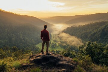 Fototapeta premium Man in Red Shirt Standing on Mountain Overlooking Green Valley and Misty Forest at Sunrise, with Orange Sky and Trees, High-Resolution Landscape