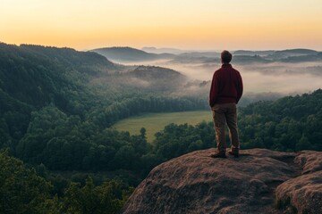 Fototapeta premium Man in Red Shirt Standing on Mountain Overlooking Green Valley and Misty Forest at Sunrise, with Orange Sky and Trees, High-Resolution Landscape