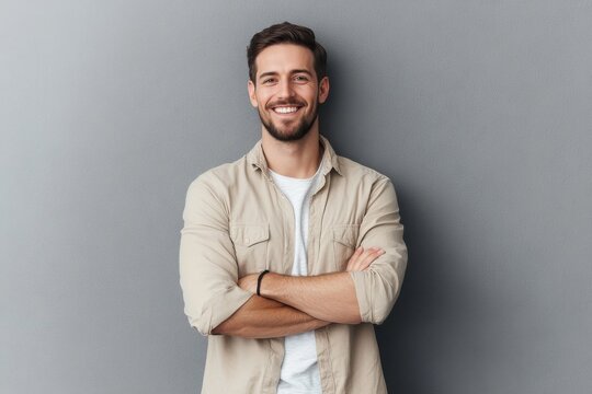 Portrait of a smiling young man in a beige shirt, standing with arms crossed, looking casually at the camera against a gray background.