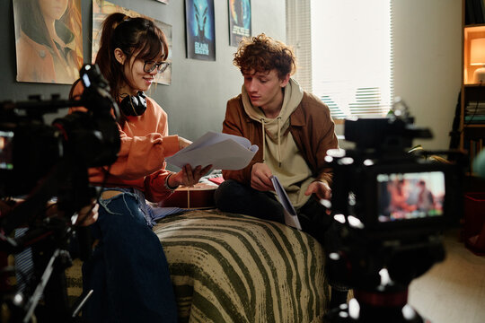 Two young individuals collaborating on a film project in a cozy room filled with cameras and movie posters, one holding script papers while the other listens attentively