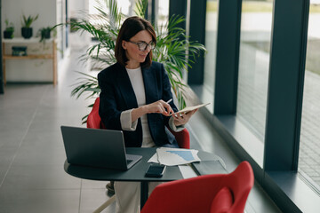A businesswoman uses a tablet and laptop in her office, focusing on work tasks with dedication
