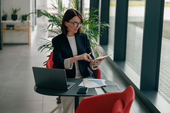 A businesswoman uses a tablet and laptop in her office, focusing on work tasks with dedication
