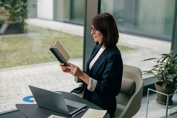 A focused businesswoman is reading a book in a modern office surrounded by a laptop and a notebook
