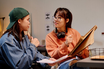 Two Asian women engaging in animated discussion while planning an art project in cozy room holding art materials and wearing casual clothes