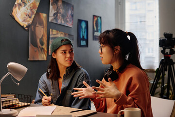 Two Asian women engaged in discussing ideas at cozy indoor workspace with posters on wall and a camera in background