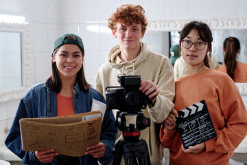 Portrait of three young people holding film equipment and smiling warmly at camera in studio...