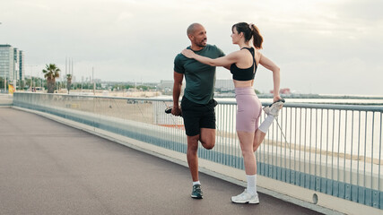 A multi-ethnic couple of young people training together in summer, male and female athletes doing muscles stretching, exercises by the sea, sporty people diverse, man and woman training