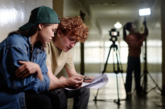 Two young individuals sitting together and reviewing script in a film production setting with a tripod and lighting equipment in background