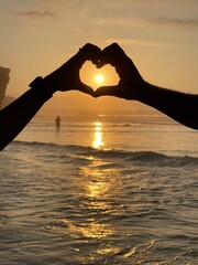 A couple forms a heart with their hands in front of the sunset by the sea. Partnership and love concept. Silhouette of hands in the shape of a heart with sunset in the middle and beach background.