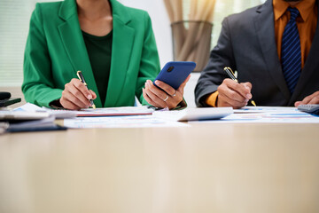 Fototapeta premium Asian business team engages in a collaborative meeting, financial charts at a desk. Dressed in formal suits, they brainstorm creative ideas to enhance company performance and team cohesion.