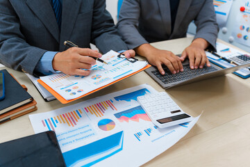 Asian business team engages in a collaborative meeting, financial charts at a desk. Dressed in formal suits, they brainstorm creative ideas to enhance company performance and team cohesion.