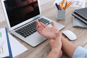 Man suffering from pain in wrist while working on laptop at table indoors, closeup. Carpal tunnel...