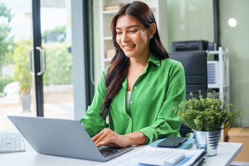An Asian businesswoman sits at the table in her office, working as an accountant. She uses a...