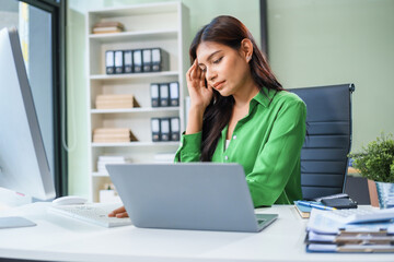 An Asian businesswoman sits at her office table, stressed and with a headache from hard work. Prolonged sitting leads to muscle weakness and the negative effects of office syndrome.