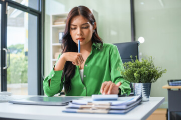 Fototapeta premium An Asian businesswoman sits at her office table, stressed and with a headache from hard work. Prolonged sitting leads to muscle weakness and the negative effects of office syndrome.