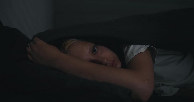 Portrait of a blonde girl in a white T-shirt who sharply falls her head in bed and covers herself with a blanket at night