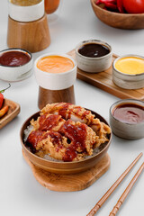A rice bowl topped with crispy chicken and drizzled with spicy red sauce, accompanied by a variety of sauces around it.
