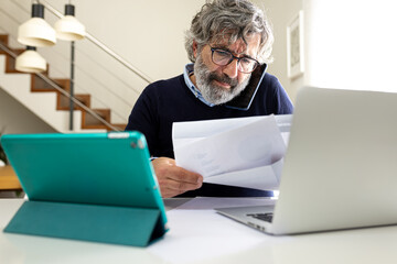 Stressed mature man working with laptop talking on the phone at home office feeling overwhelmed.