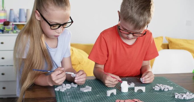 Concentrated little children build houses of toy clay bricks at table. Brother and sister use ceramic blocks making architecture at home