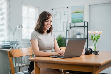 Smiling young woman using a laptop at a wooden desk in a bright home office with modern decor and tulips.