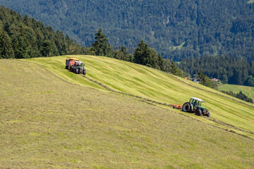 Two tractors (hay tedder and trailor) in use during hay harvest on a mountain meadow in Tyrol,...