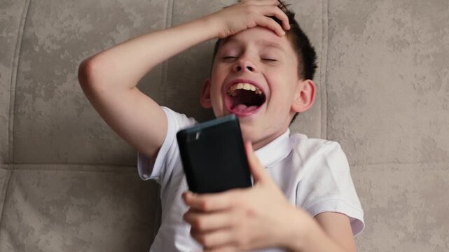 Caucasian boy 10-11 years old looks at the screen of a smartphone or gadget and laughs from the content or meme he sees, funny media on the phone for a child
