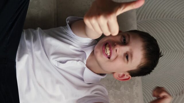 portrait of caucasian cheerful boy 10-12 years old showing with hands gesture like or cool sitting on sofa and looking at camera, vertical video