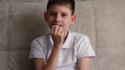 school age boy biting nails, caucasian boy sitting on sofa biting nails and looking at camera, sign of doubt and insecurity