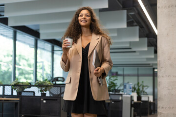 Businesswoman walking along the office corridor with documents and paper cup.