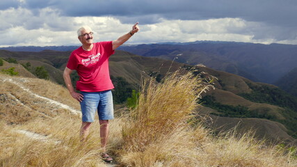 aktiver Senior wandert im Landesinneren von Sumba in bergiger Landschaft und zeigt in Windrichtung...