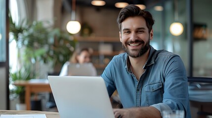 Obraz premium Portrait of male employee with laptop and smiling at camera in office interior, successful businessman in classic spectacles posing at desktop with laptop computer